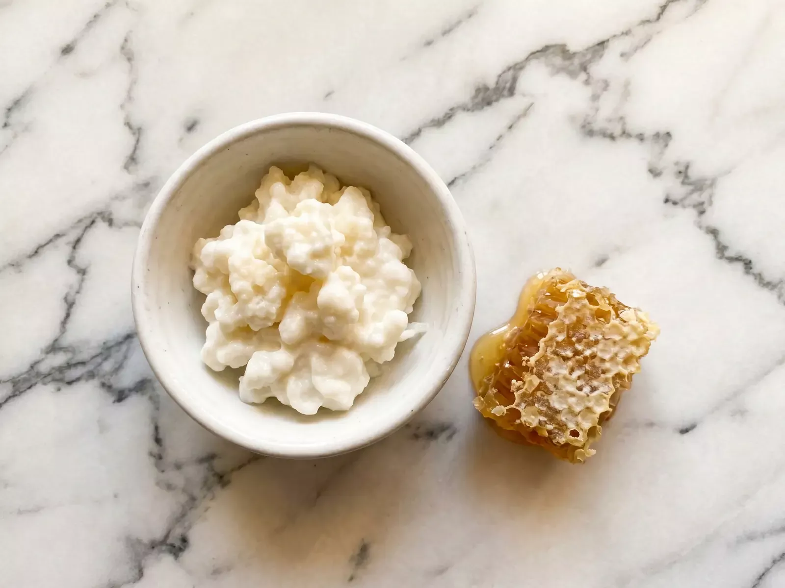 Cuenco de kefir natural com panal de mel ao lado, sobre m&aacute;rmore com luz quente cenital.